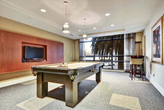 Room with a pool table and seating area, featuring large windows and a wall-mounted TV.