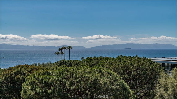 Panoramic view of the ocean and distant islands with trees and a rooftop in the foreground.