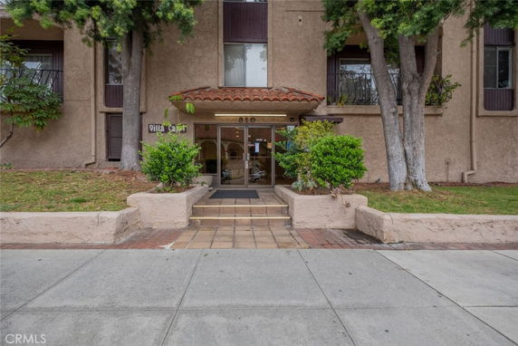 Front view of a building entrance with trees and shrubbery, featuring a tiled walkway.