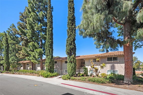 Front view of a two-story residential building with multiple units and a tiled roof, surrounded by tall trees.