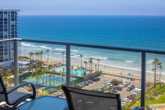 Panoramic view of ocean, beach, swimming pool, and tennis courts seen from a balcony.