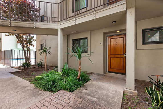 Partial view of an apartment entrance with a wooden door, surrounded by plants and a walkway, near a fenced area.