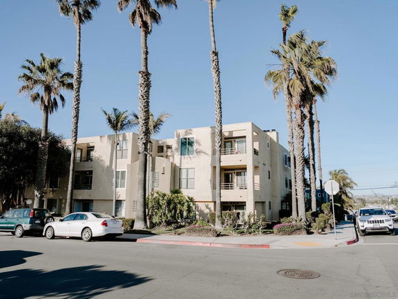 Front view of a three-story apartment building with balconies and surrounded by palm trees.