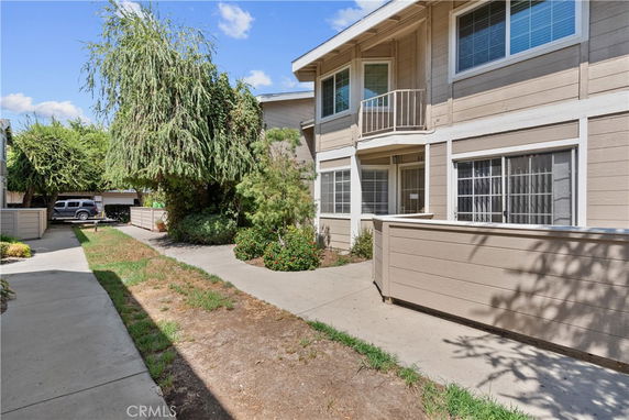 Exterior view of a two-story residential building with beige siding and white trim, featuring a small fenced patio and adjacent walkway.