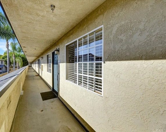 Exterior view of a long, narrow corridor with beige textured walls and windows, leading to individual apartment entrances.