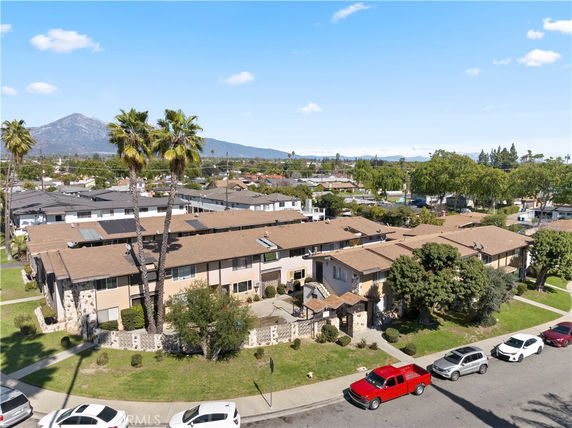Elevated view of a two-story apartment complex with brown roofing and surrounding greenery, located in a suburban area with mountains in the background.