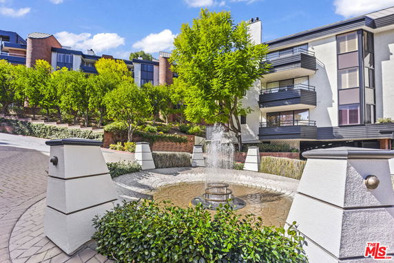 Front view of a modern multi-story residential building with balconies and a circular fountain in the foreground.