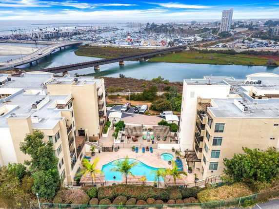 Aerial view of two modern apartment buildings with a swimming pool area in between, overlooking a river and a marina in the background.