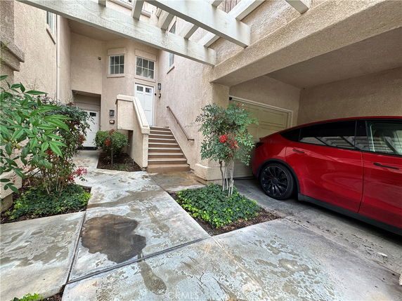 Covered parking area with a red car next to a stairway leading to the entrance of a building.