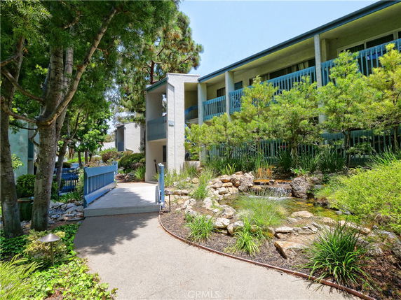 Front view of a multi-story building with blue railings and a small landscaped area with water feature.
