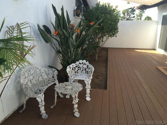 Outdoor patio area with decorative white chairs and a small table, next to plants and a wooden deck.