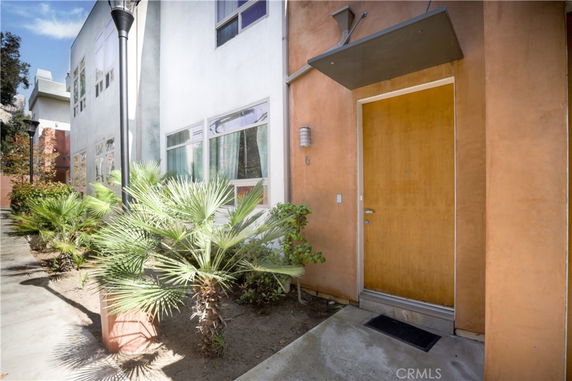 Front view of a modern multi-story building with a wooden entrance door and small plants alongside the pathway.