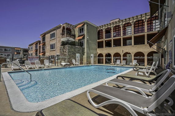 Outdoor swimming pool area with lounge chairs surrounded by multi-story residential buildings.