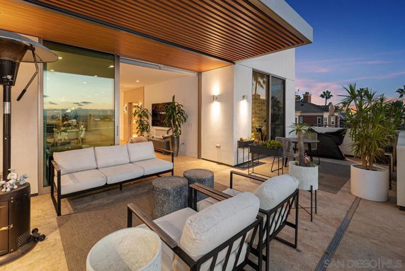 Rooftop seating area with modern furniture and decorative plants, adjacent to a sliding glass door leading to the interior.