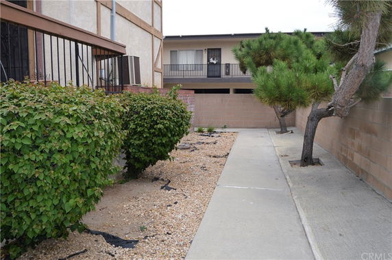 Side view of a two-story building with balconies and trimmed bushes alongside a concrete pathway.