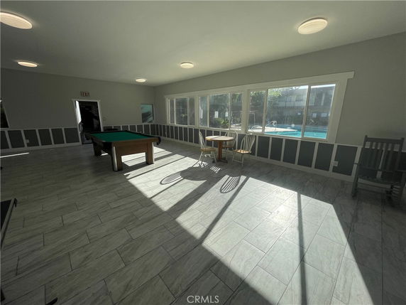 Room with a pool table and seating area, featuring large windows overlooking an outdoor pool.
