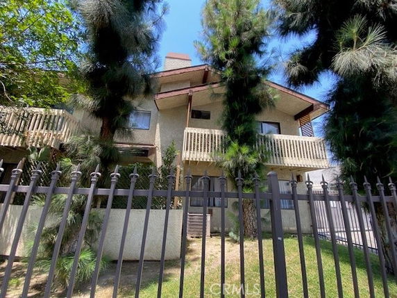 Front view of a two-story house with balconies, surrounded by tall trees and a metal fence.