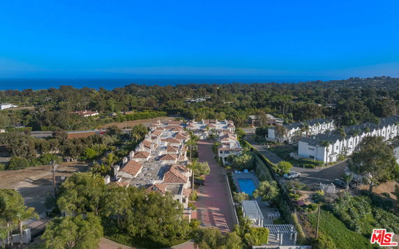 Aerial view of a residential complex with red-tiled roofs, surrounded by greenery and set against a backdrop of a blue sky and distant hills.