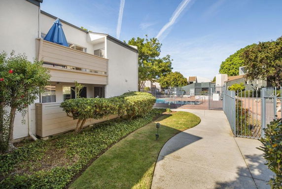 View of a residential building with a balcony and pathway leading to a fenced swimming pool area.