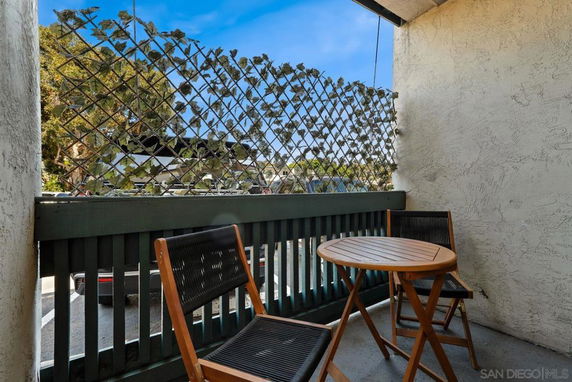 Small balcony with chairs and a table, featuring a lattice fence with plants and a view of parked cars.