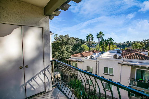 View from a balcony overlooking adjacent buildings with terracotta roofs and a backdrop of trees and palm trees.
