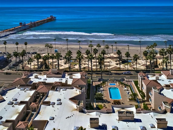 Panoramic view of beachfront buildings with a swimming pool and ocean pier in the background.