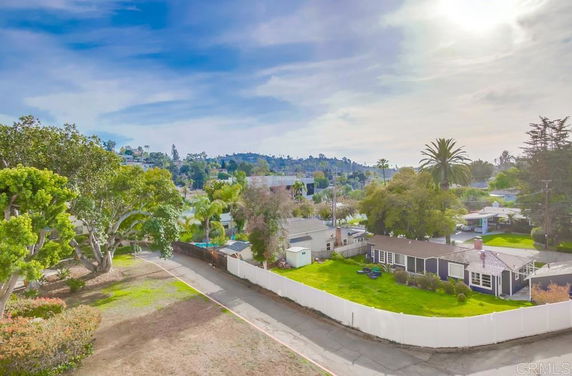 Aerial view of a neighborhood with houses, trees, and a distant hillside under a partly cloudy sky.