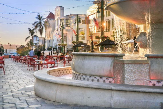 Outdoor area with a large fountain, seating, and buildings with flags.