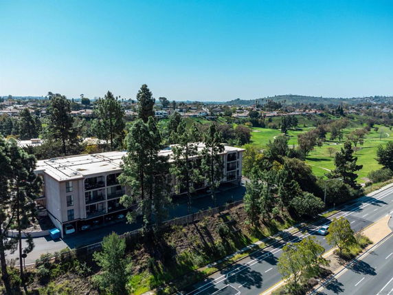 Aerial view of a multi-story residential building surrounded by trees, with a wide road and expansive landscape in the background.