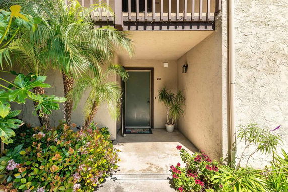 Front view of a residential entrance with a door and a small balcony above, surrounded by plants.
