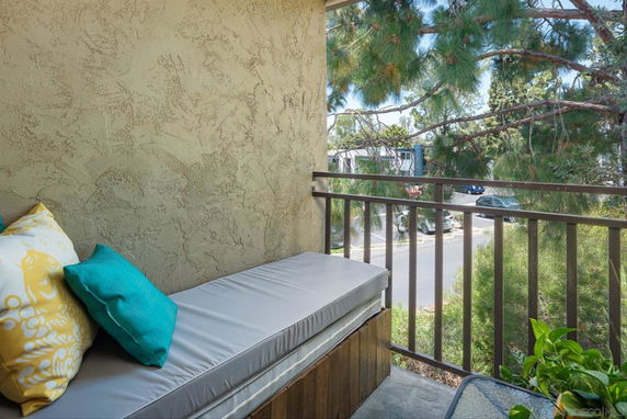 View from a balcony with outdoor seating and a railing, overlooking trees and a parking area.