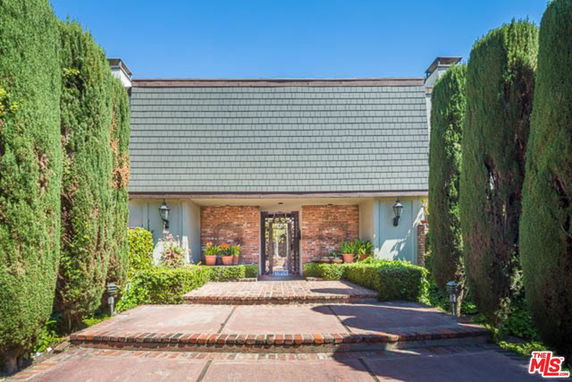 Front view of a house with a shingled roof and brick facade, flanked by tall bushes.