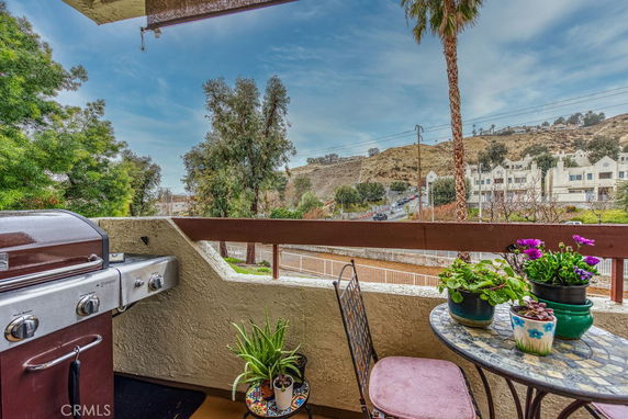 Outdoor balcony with a grill, small table, chairs, and potted plants overlooking a hillside and residential area.