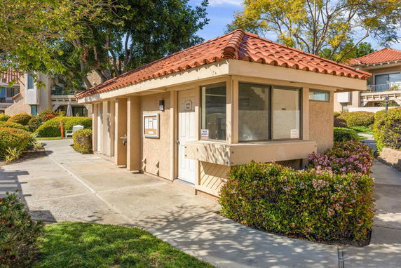 Front view of a small building with a red tile roof and beige exterior, surrounded by shrubs.