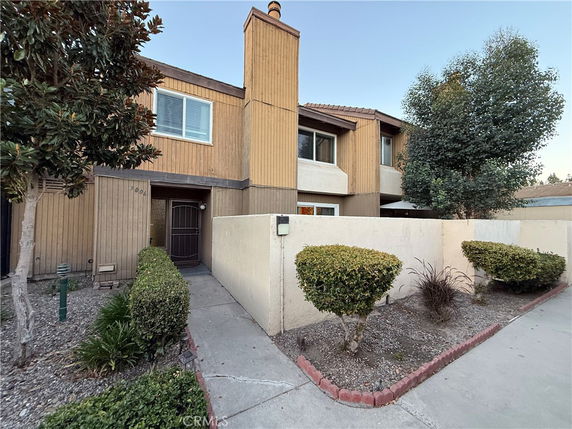 Front view of a two-story house with wood paneling and a reinforced door entrance, surrounded by a low wall and shrubbery.