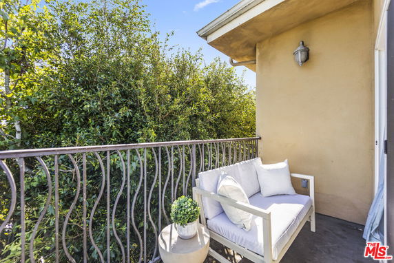 Small outdoor balcony with seating and a decorative plant, adjacent to the building wall, with greenery in the background.