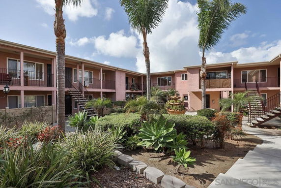 View of a two-story apartment building with balconies and exterior staircases, surrounded by landscaped garden areas.