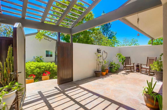 Outdoor patio area with wooden chairs and potted plants, partially covered by a pergola.