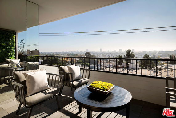 Panoramic view from a balcony with seating and a table overlooking a cityscape.