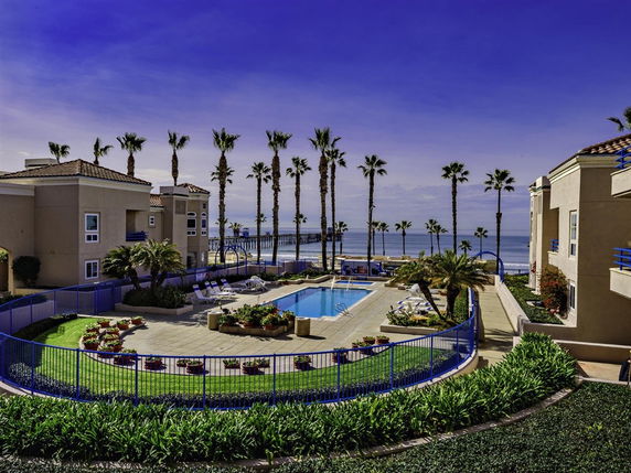 Outdoor pool area surrounded by buildings and palm trees, with a view of the ocean.
