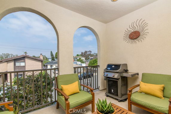 Balcony with arched openings, outdoor seating, and a grill, overlooking a residential area.