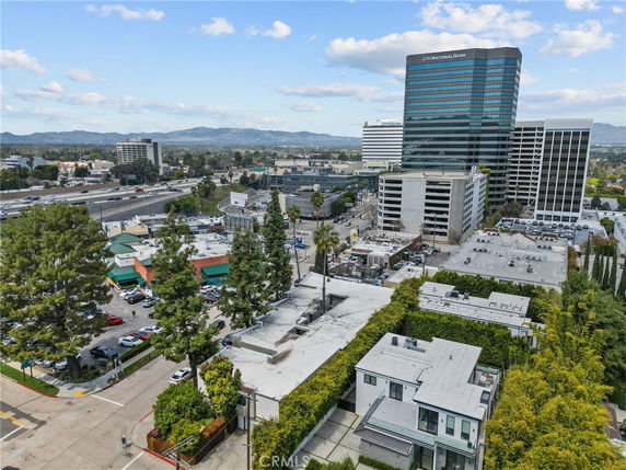 Aerial view of a cityscape with multiple buildings, including a multi-story modern building and smaller commercial structures, surrounded by greenery and roads.