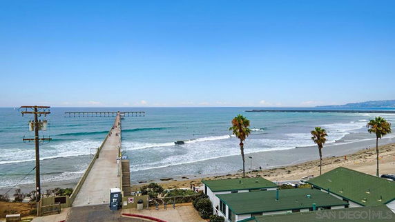 Panoramic view of a beach with a pier extending into the ocean and green-roofed buildings in the foreground.
