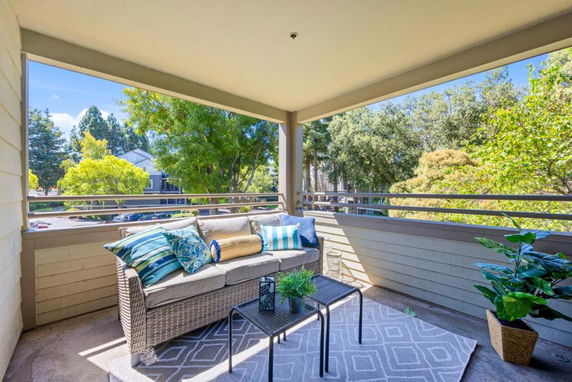 Covered balcony with sofa and table, offering a view of trees and surrounding area.