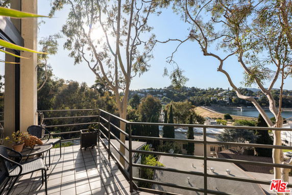 Balcony view overlooking distant hills and a body of water, with trees and rooftops in the foreground.