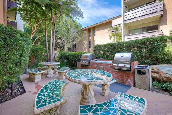 Outdoor area with mosaic tables, benches, and barbecue grills between buildings.