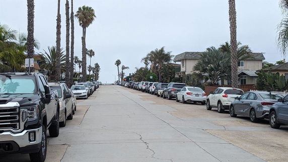 Outdoor street parking area lined with cars on both sides and residential buildings in the background.