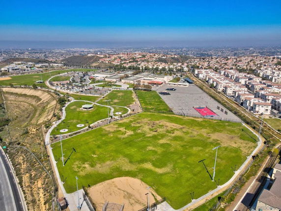 Aerial view of a large grassy park surrounded by housing and roads, featuring walking paths and parking areas.