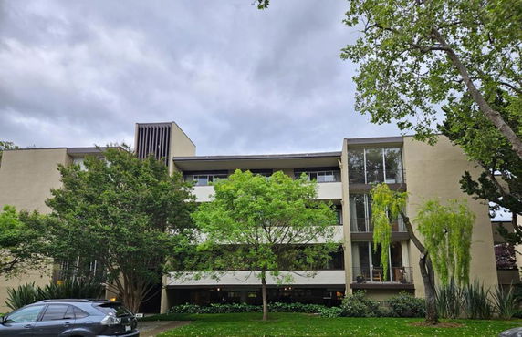 Front view of a multi-story apartment building with large windows and surrounded by trees.