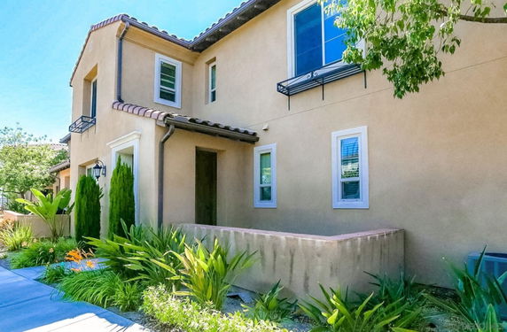 Front view of a two-story house with beige stucco walls and small windows, surrounded by plants.